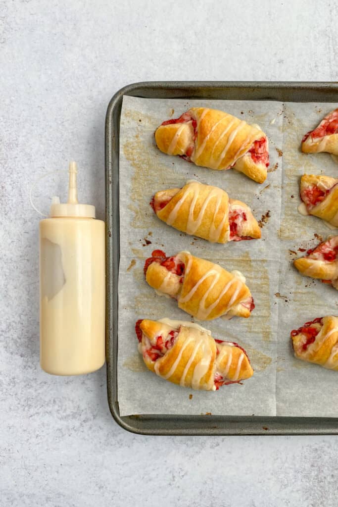 Baking sheet of crescent rolls with glaze on top of each roll, sitting next to a squeeze bottle full of the glaze.
