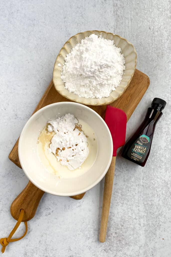 Mixing bowl with ingredients in it needed to make vanilla glaze, which is next to a bowl of powdered sugar and a bottle of vanilla extract.