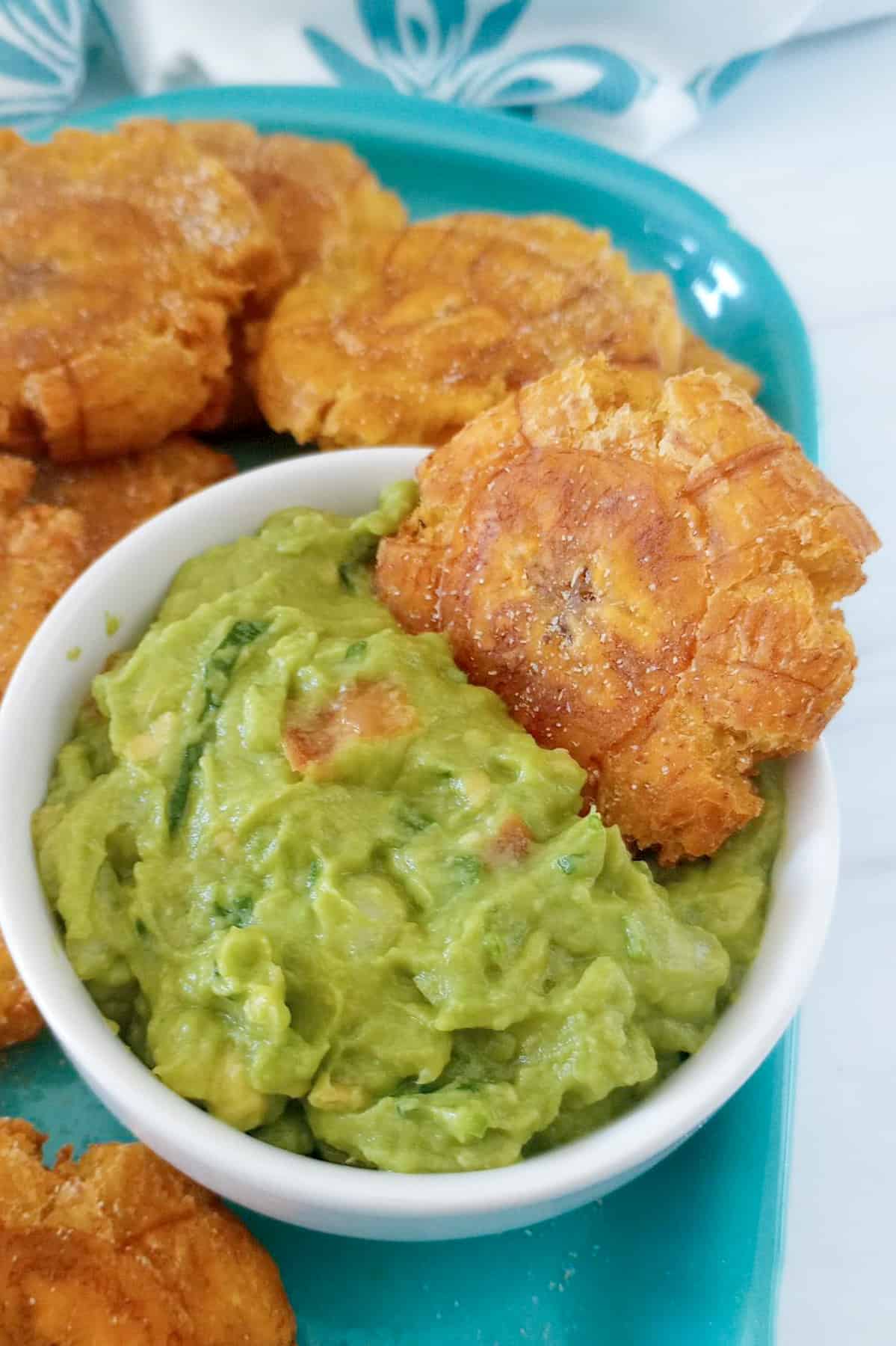 Fried green plantain being dipped in a bowl of guacamole that's sitting on a platter full of tostones.