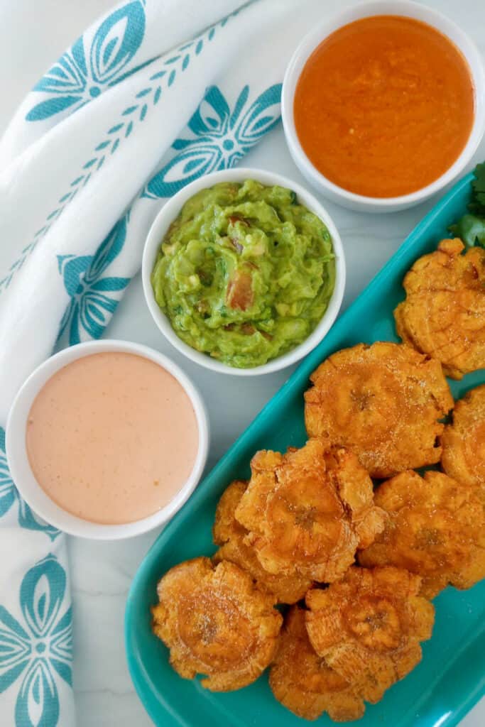 Tray of fried green plantains next to three dipping sauces, including a ketchup and mayo sauce, and guacamole.