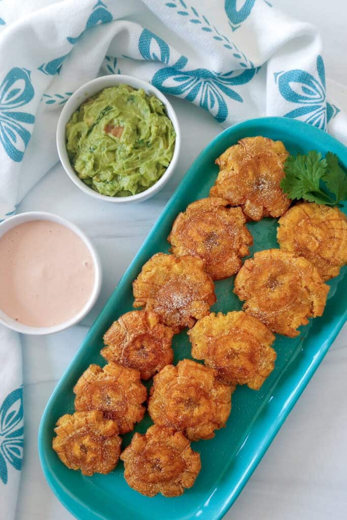 Tostones on a plate next to a bowl of mayo-ketchup dipping sauce and a bowl of guacamole.