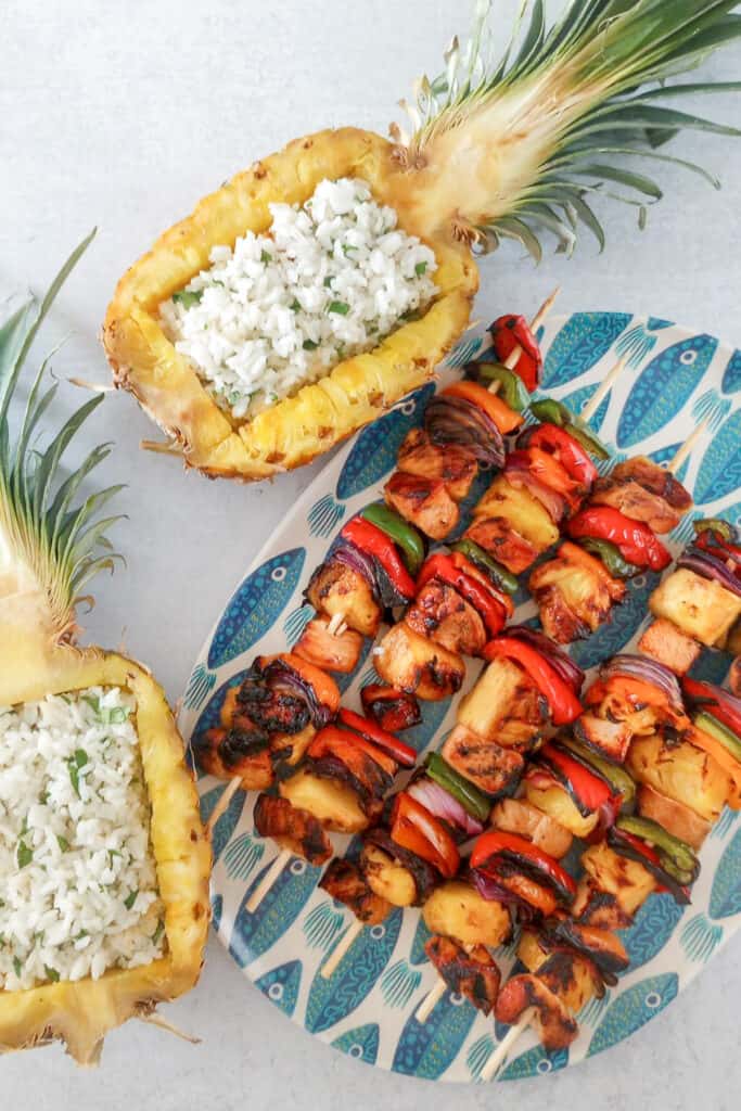 Chicken Kabobs arranged on a blue fish plate next to a hallowed out, fresh pineapple that is filled with jasmine rice. 