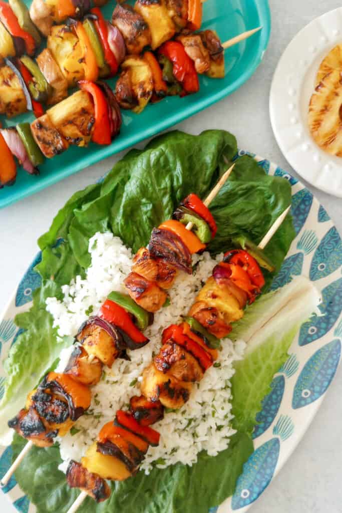 Pineapple Chicken Skewers arranged on jasmine rice on top of romaine lettuce. More Kebabs are laid next to a blue plate of pineapple kabobs and a white plate with charred pineapple assembled to the left. 