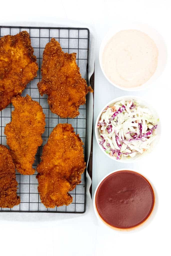 Cooked Nashville Hot Chicken next to Cole slaw Cajun remoulade sauce and Nashville hot sauce.