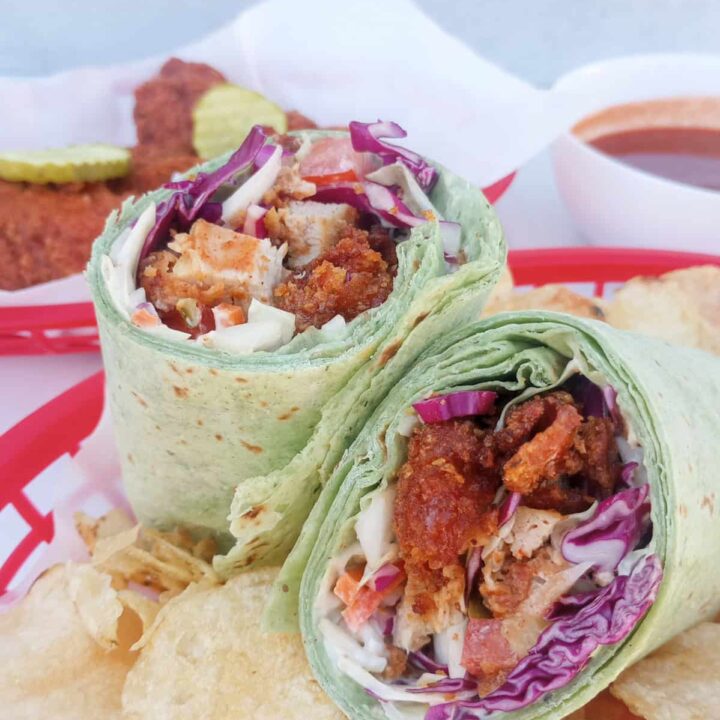 Nashville hot chicken wrap in a red basket lined with red and white checkered paper. In the background are crispy, spicy, Nashville chicken with dill pickles on top and a bowl of Nashville hot sauce in a white bowl.