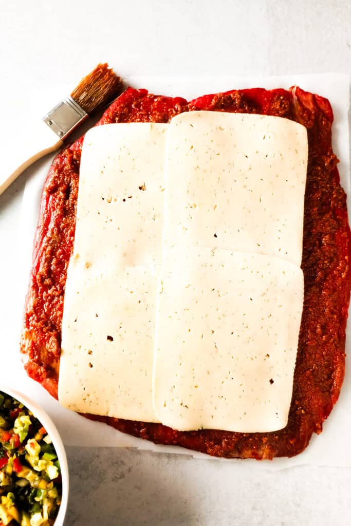 Flank steak coated in red pesto sauce topped with cheese slices next to a brown basting brush and a white bowl of mango avocado salsa. 