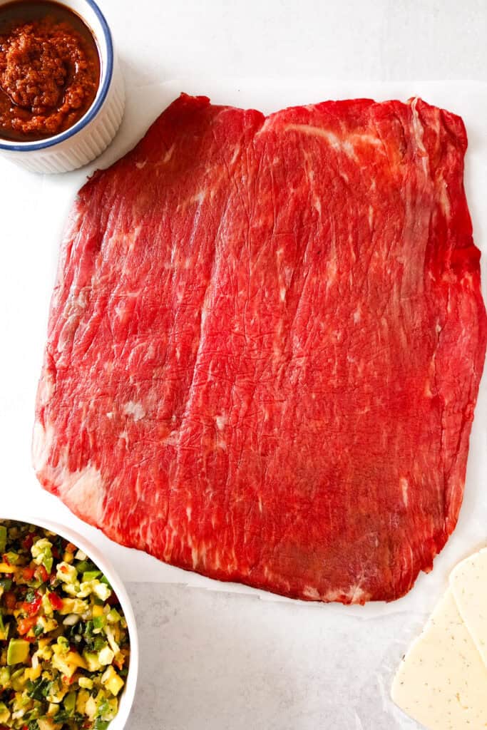 Flattened flank steak on white parchment paper next to a white bowl filled with mango avocado salsa, red pesto sauce and Havarti dill cheese slices.