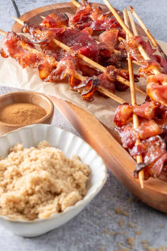  Bacon on sticks arranged on a wood serving board next to a bowl of brown sugar and bowl of cinnamon.