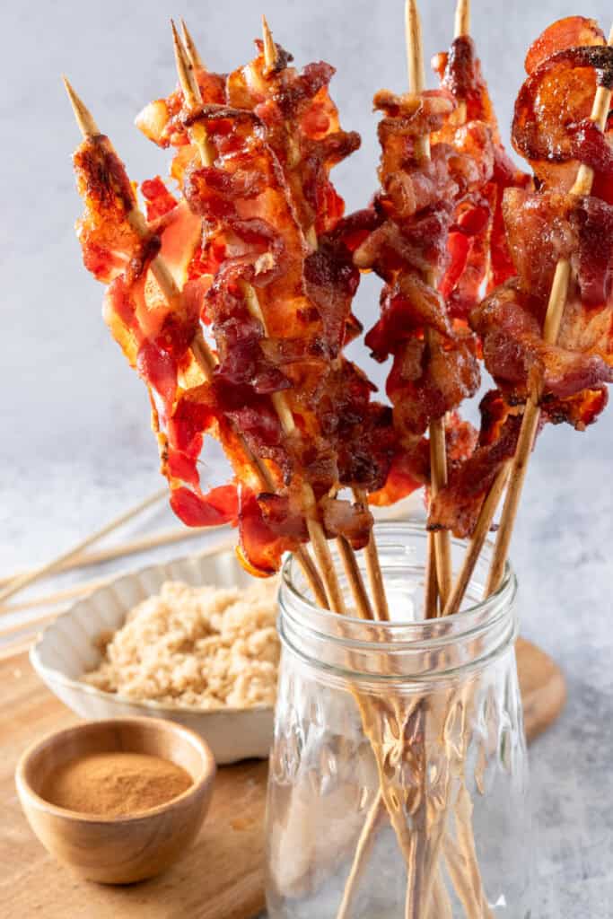 Candied bacon skewers standing up in a glass jar next to a bowl of brown sugar and ground cinnamon.