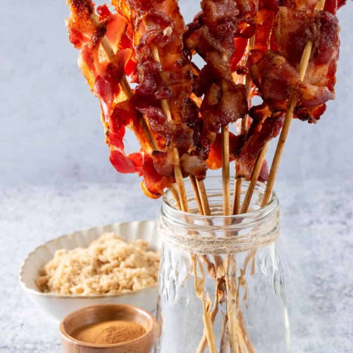 Candied bacon on skewers in a jar next to a bowl of brown sugar and cinnamon for the glaze.