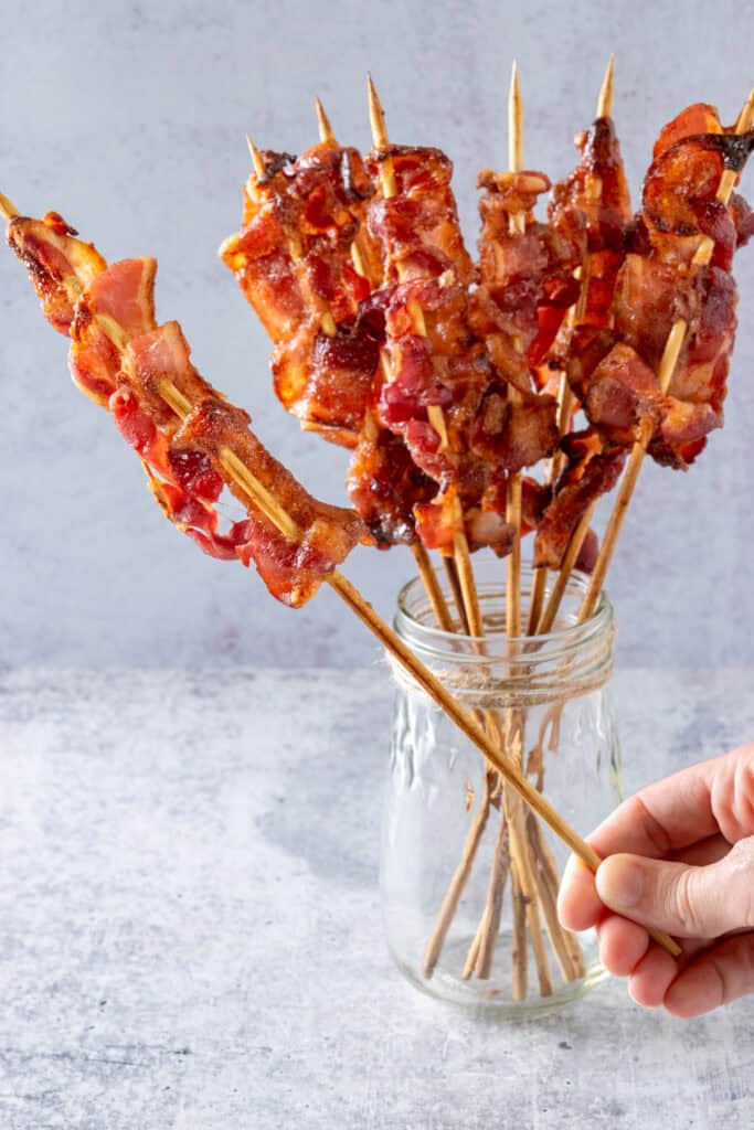 Jar holding brown sugar glazed bacon that is skewered onto sticks, with a hand holding one of the candied bacon skewers out of the jar.