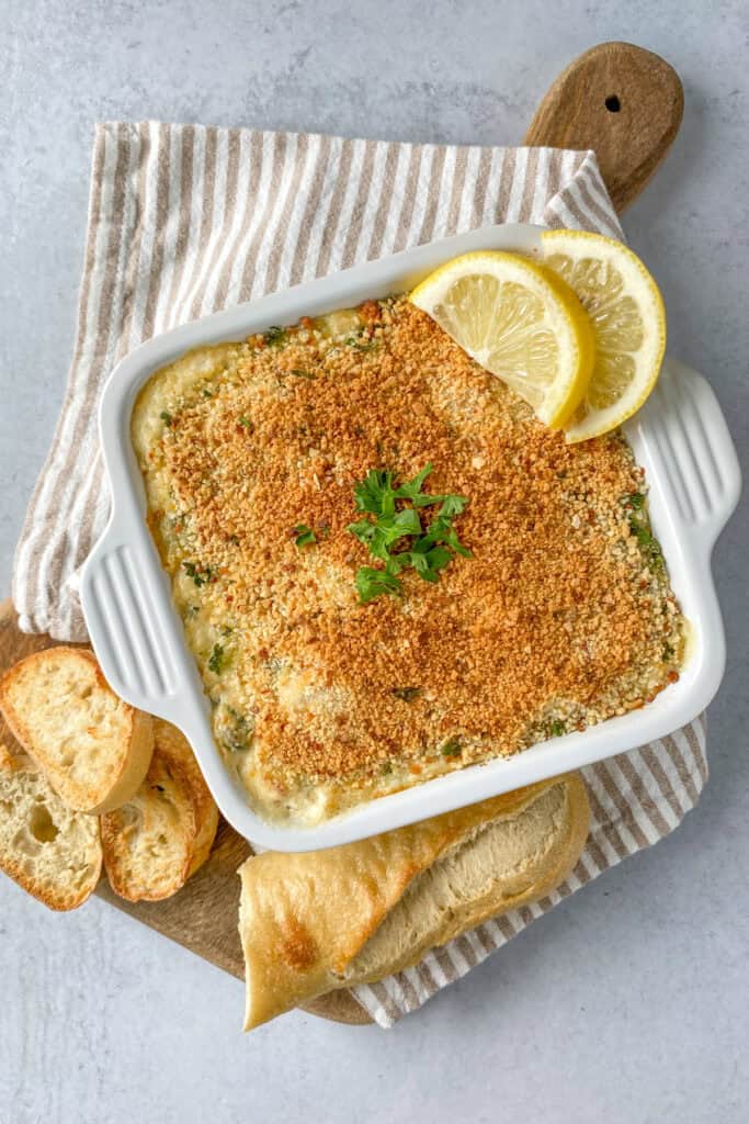 Crab and spinach dip in a square baking pan, topped with bread crumbs and garnished with lemon wedges and parsley.