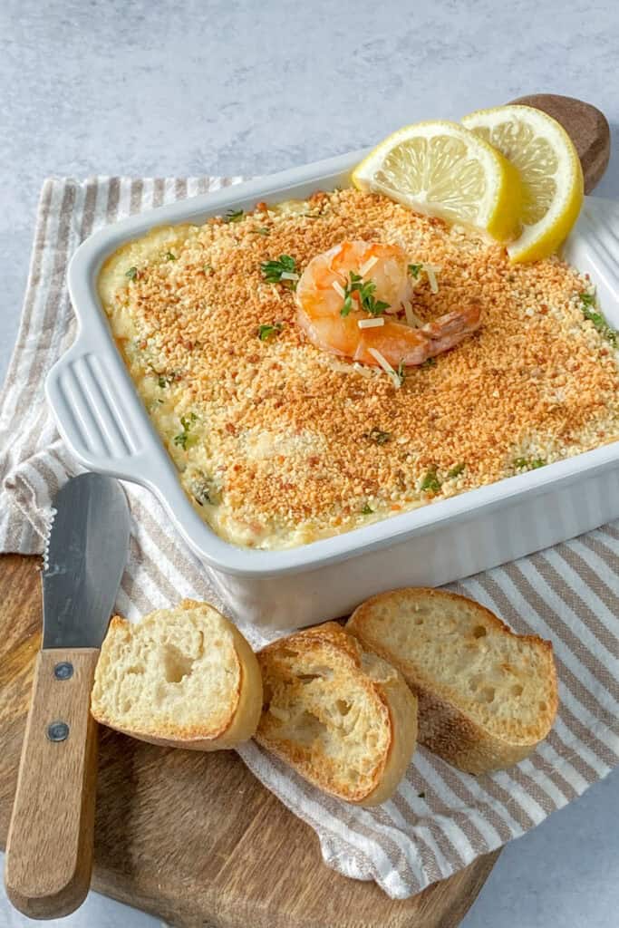 Seafood dip fresh out of the oven topped with browned Parmesan crusted breadcrumbs, shrimp and lemon wedges placed on the side. Served on a brown chopping board layered with a white and tan cloth with warmed baguette bread. 