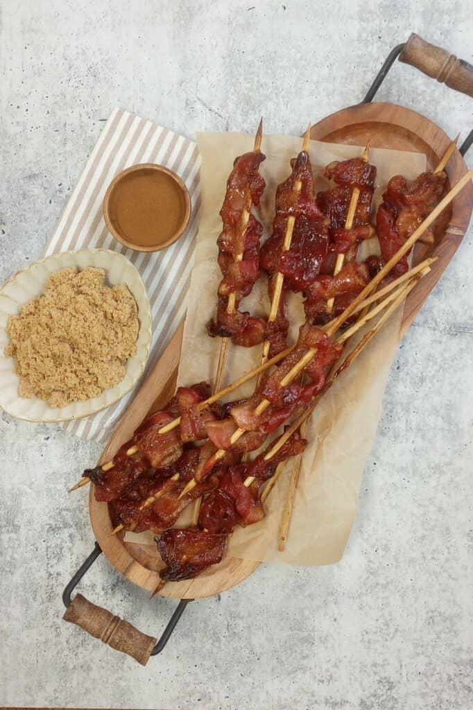 Bacon on a serving tray next to a white and brown towel, brown sugar, and cinnamon. 