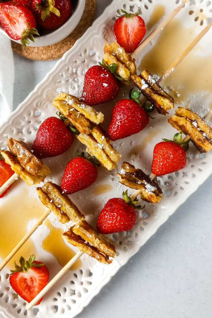 Breakfast kebabs up close served on a white serving plate. A white towel is angled around the plate for dimension and strawberries are sliced and layered on the white plate.