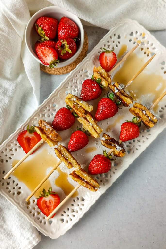 French toast kabobs are made with sandwich bread and strawberries. Breakfast kebabs are angled in threes on a white serving plate with sliced strawberries layered on the plate. Maple syrup is drizzled on the white serving plate for dimension.