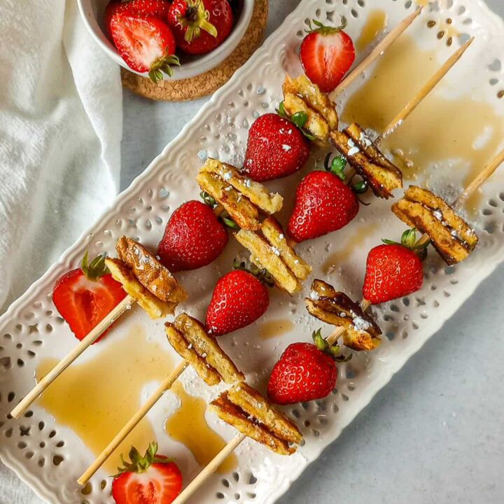 French toast kabobs are made with sandwich bread and strawberries. Breakfast kebabs are angled in threes on a white serving plate with sliced strawberries layered on the plate. Maple syrup is drizzled on the white serving plate for dimension.