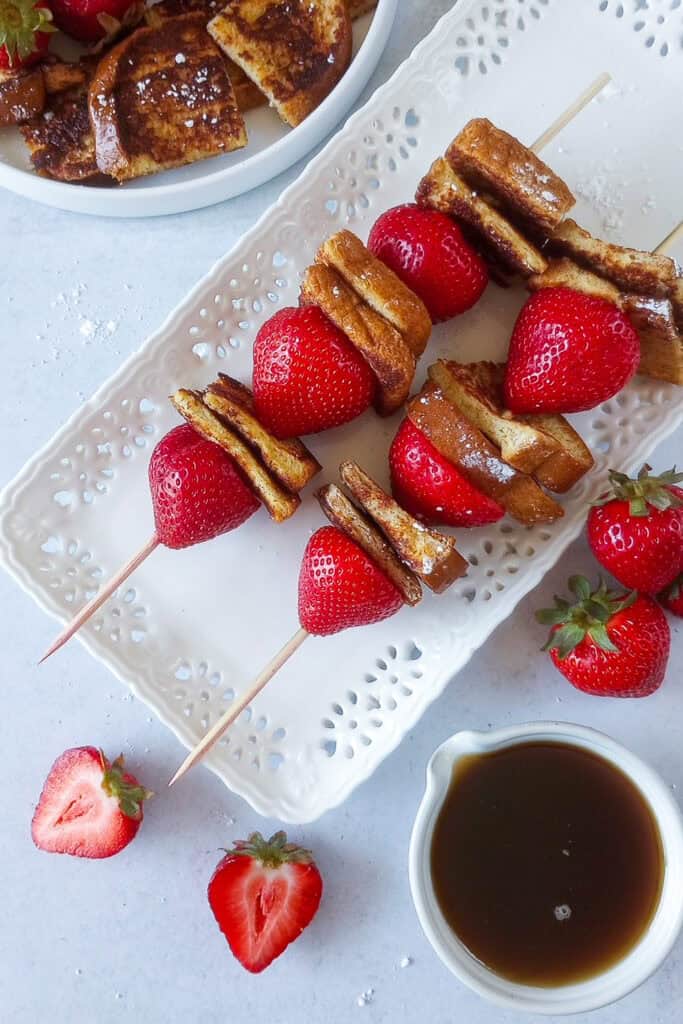 Up close picture of breakfast kabobs next to syrup. Sliced strawberries are arranged around background and powdered sugar is sprinkled over the background for a little dimension. 