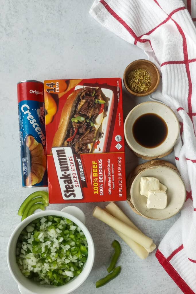 Diced green pepper and yellow onion in a white bowl arranged next to sliced green bell peppers. String cheese is arranged in thirds angled next to the bowl of diced peppers and onions.  A red and white checkered towel is scrunched up on the right side and follows the three bowels, which contain sliced butter, Worcestershire sauce, and Montreal steak seasonings. Canned crescent rolls and sliced steak-umms are placed in the middle of the photo graph next to the three bowels of ingredients. 