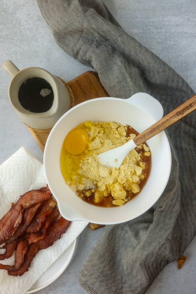 Jiffy mix, egg, maple syrup, milk, combined in a white mixing bowl with a stiring spoon ready to be mixed. White bowl is on a brown chopping board arranged next to cooked bacon, maple syrup, and a bunched up grey and black towel. 