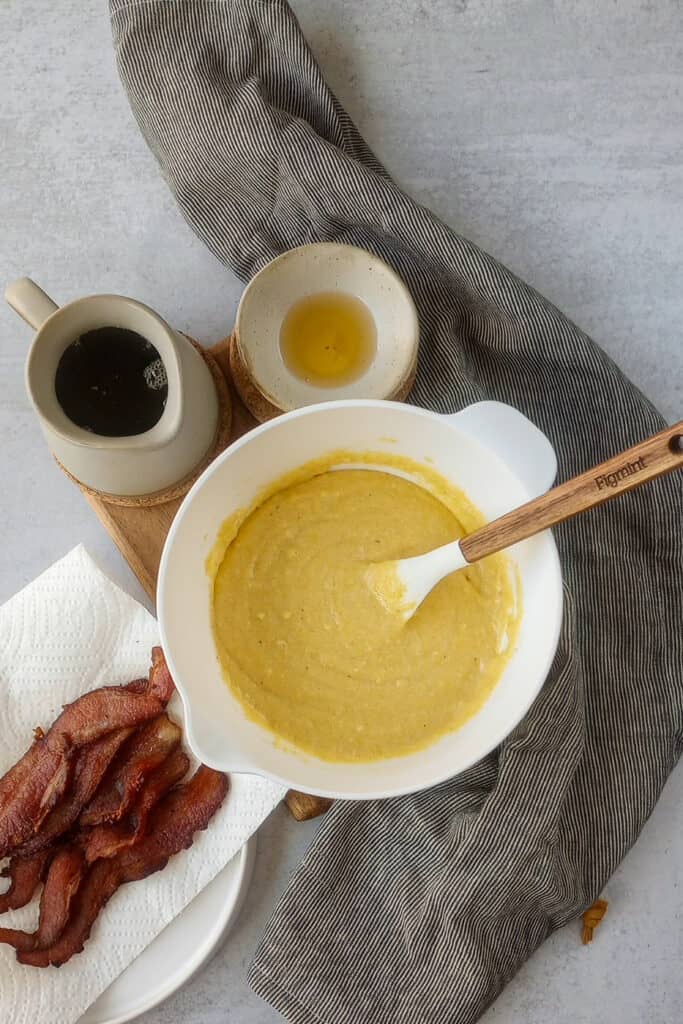 Cornbread mix arranged next to maple syrup, bacon, grey and black towel, and bacon grease. 