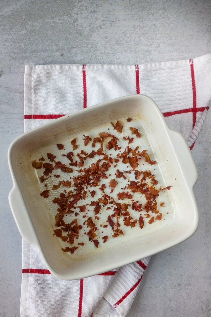 A white baking dish coated with bacon drippings and half crumbled bacon is added to the bottom of the baking dish for lots of bacon flavor. 