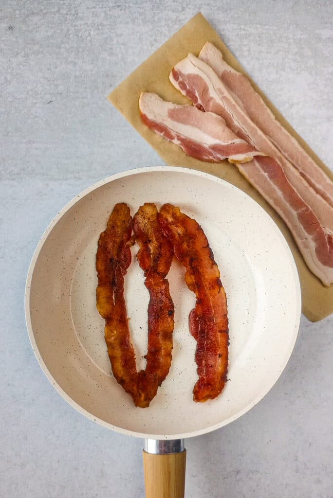 Cooked bacon in a white skillet with a light tan handle. Bacon slices are arranged in the top right corner above the pan for dimension. 