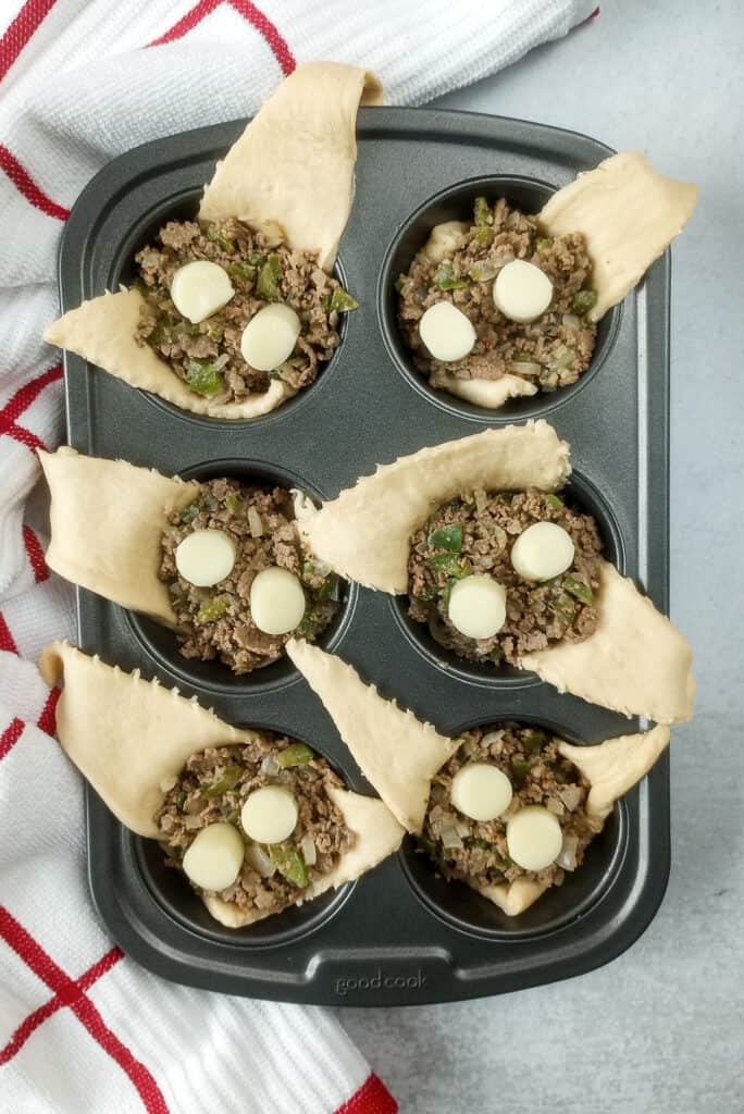 Muffin tin arranged next to a red and white checkered towel. Crescent rolls are topped with the last layer of sliced string cheese and ready to be closed shut. 