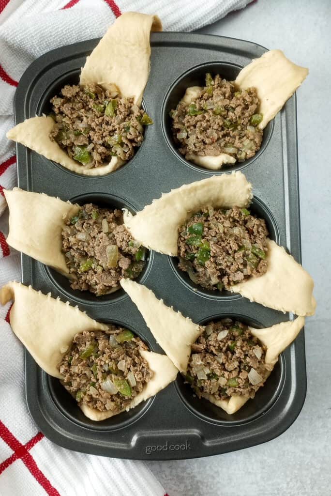 Muffin tin laid next to a red and white checkered towel. Crescent rolls are stuffed with meat and cheese. 