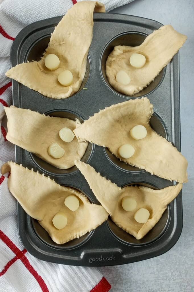 Muffin tin laid out next to a white and red checkered towel with crescent rolls filled with sliced string cheese. 