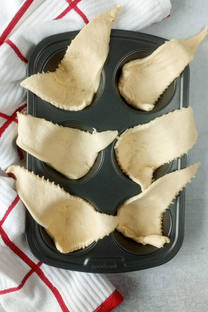 In a greased muffin tin, crescent rolls are arranged and muffin tin is arranged next to a red and white checkered towel ready to be filled with sliced string cheese. 