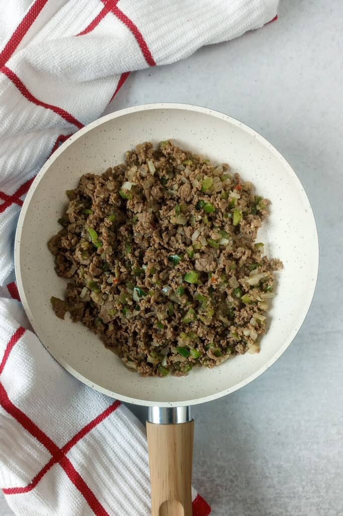 Cooked steak-umms, green pepper, and onion in a white skillet with a tan handle laid next to a red and white checkered towel. 