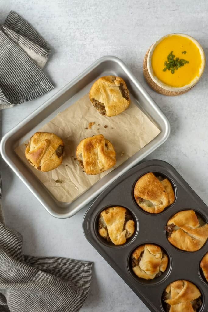 Crescent rolls fresh out of the oven in a grey muffin tin next to a silver baking sheet. A Grey and white striped towel is arranged around the left corner to add dimension in the photograph. 