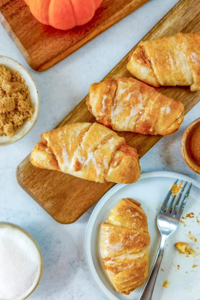 Pumpkin cream cheese crescent rolls on a brown board next to a tiny orange pumpkin. Brown sugar, pumpkin pie spice, and granulated sugar are arranged in bowls. Center stage is a pumpkin cheesecake roll on a dessert plate next to a dessert fork.