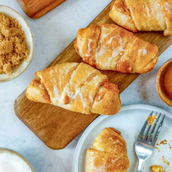 Pumpkin cream cheese crescent rolls on a brown board next to a tiny orange pumpkin. Brown sugar, pumpkin pie spice, and granulated sugar are arranged in bowls. Center stage is a pumpkin cheesecake roll on a dessert plate next to a dessert fork.