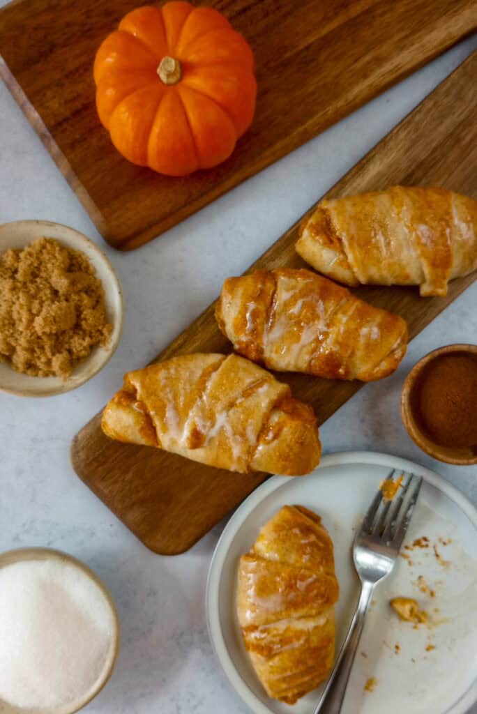 Far away picture of pumpkin crescents on a brown board next to a dessert plate with a breakfast pastry and dessert fork. Arranged around the pastries is a bowl of granulated sugar, brown sugar, pumpkin pie spice and a tiny orange pumpkin. 