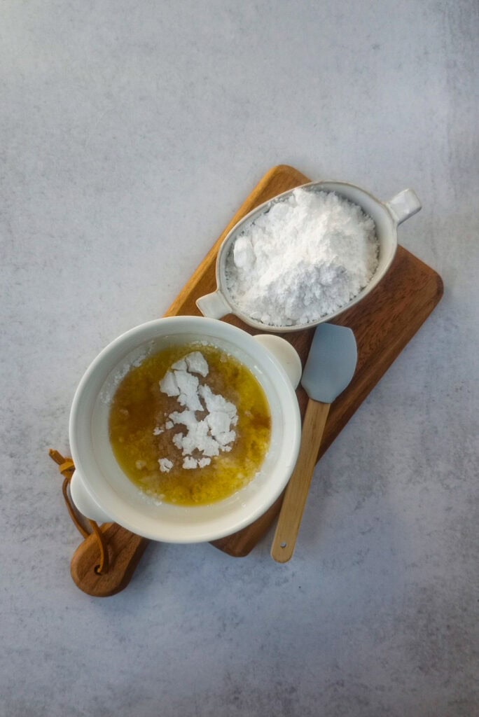 Powdered sugar, vanilla extract, melted butter in a white mixing bowl on a broad board next to a blue and tan silicone spatula.