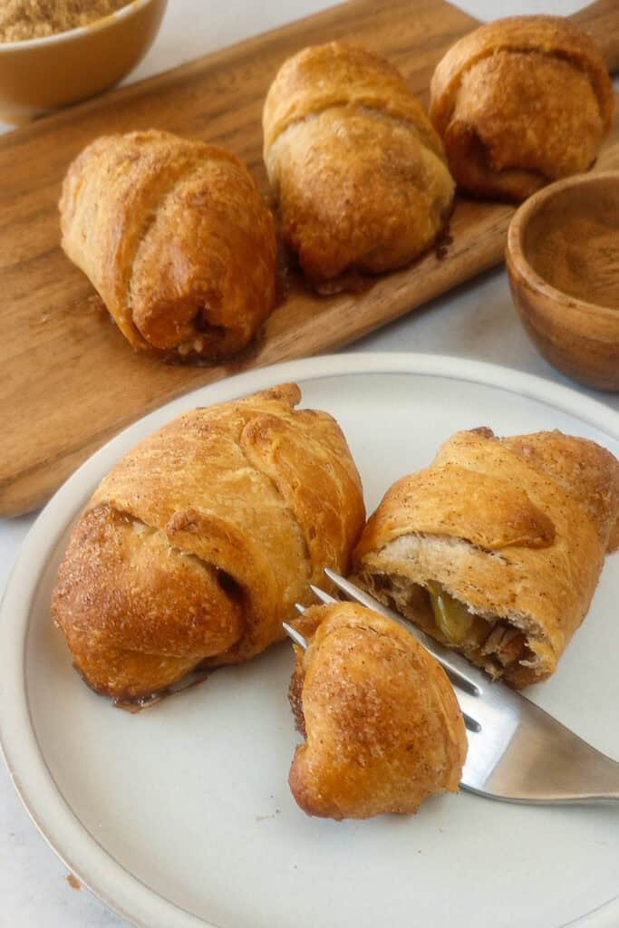 Up close picture of apple pie bites with crescent rolls on a white plate. Two apple pie bites are on the plate and one is sliced open with a dessert fork.