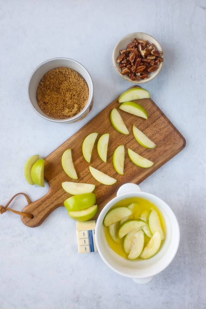 Overhead picture of sliced green apples, bowl of cinnamon sugar mixture, bowl of sliced apples in a bowl with melted butter and chopped pecans. 