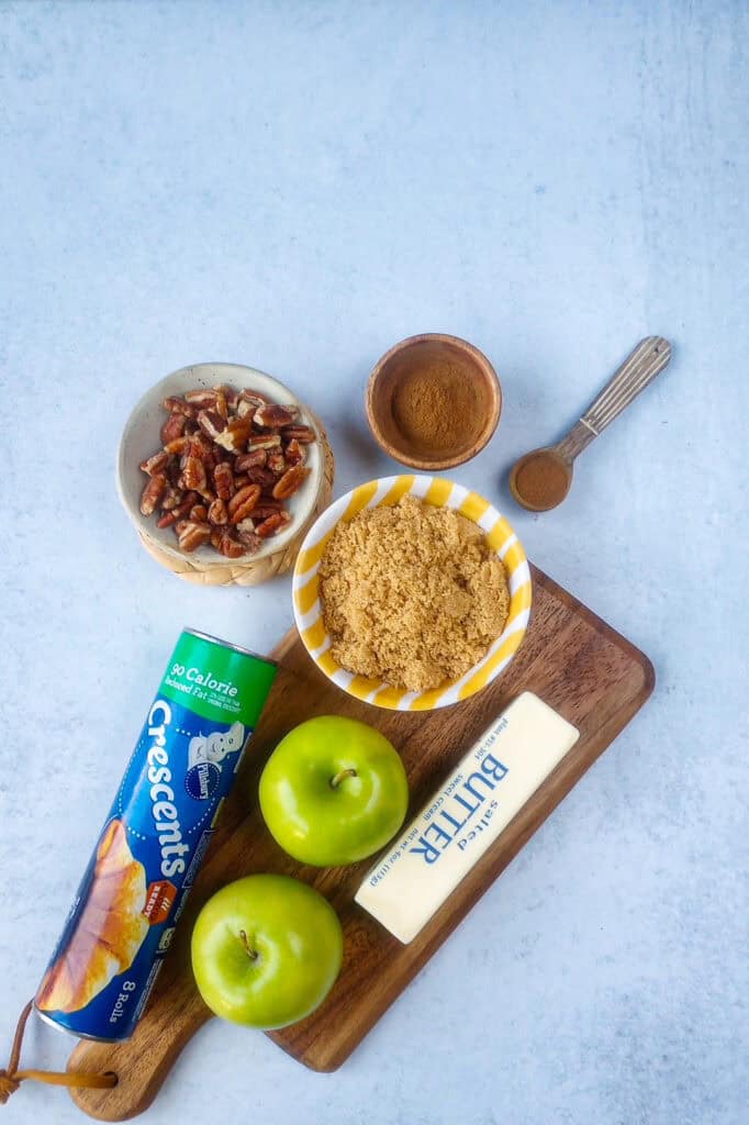 Overhead picture of all the ingredients needed to make mini apple pie bites with Pillsbury Crescent Rolls. Two green apples, 1 can of Pillsbury reduced fat crescent rolls, brown sugar, cinnamon, pecans, and salted butter. 