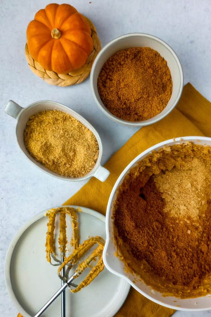 Pumpkin cream cheese filling in a white bowl topped with the last 1/2 cup of crushed graham crackers, crumbled ginger snap cookies ready to be blended. 