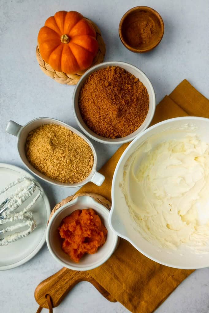 Whipped, softened, cream cheese and powdered sugar in a mixing bowl next to other ingredients; pumpkin puree, cinnamon, pumpkin pie spice, crumbled graham crackers, crumbled ginger snap cookies. 