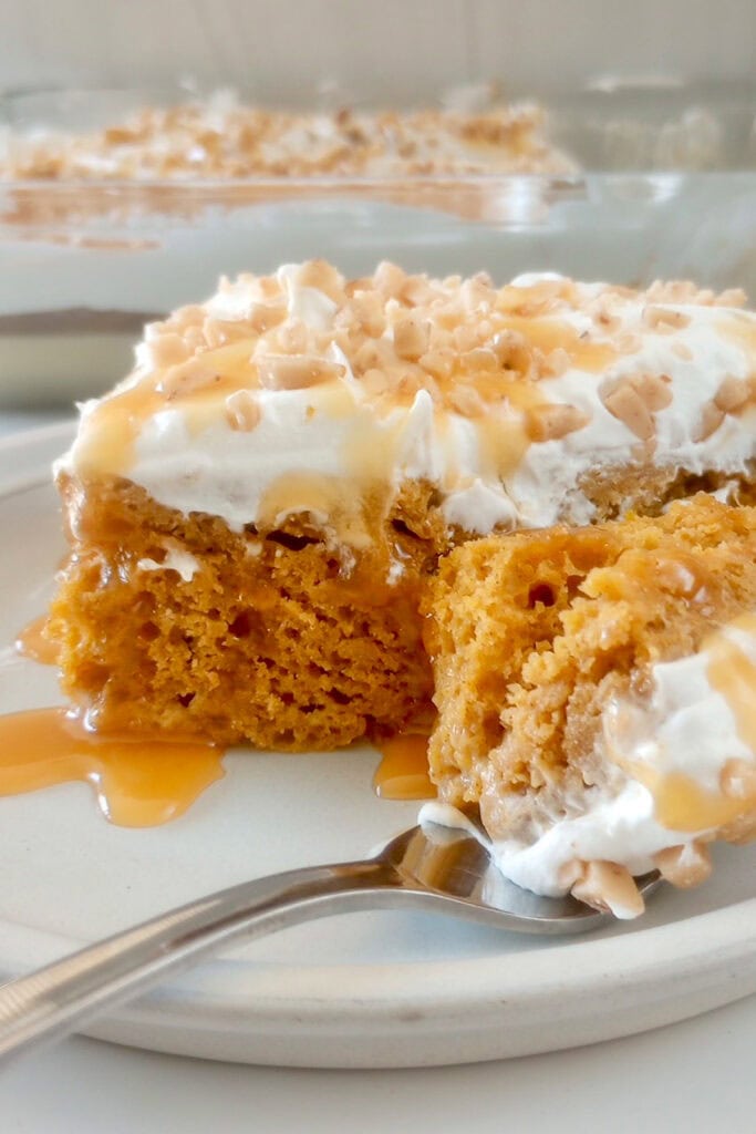 A slice of pumpkin poke cake with whipped cream, caramel and toffee topping on a plate with a fork.