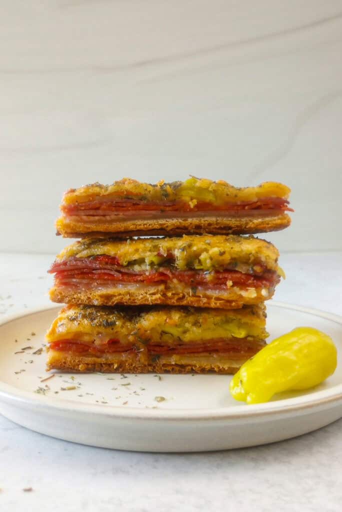 Straight on picture of antipasto squares stacked in threes on a white plate next to a whole pepperoncini. 