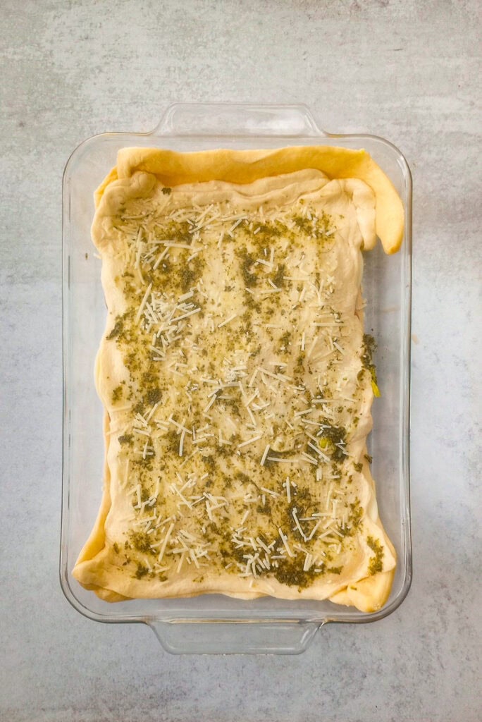 Overhead picture of top crescent roll dough seasoned with pesto sauce and seasonings, sprinkled with grated Parmesan cheese. 