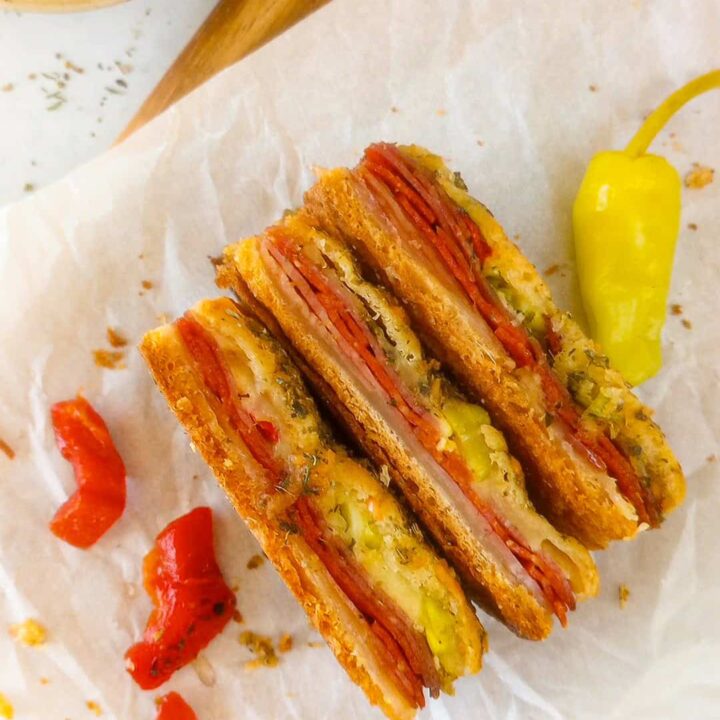 Overhead picture of antipasto squares on white parchment paper laid on the side showing meat and cheeses. Red peppers are arranged on the left side and one whole pepperoncini on the right side. Italian seasoning is in a white bowl in the upper left corner.