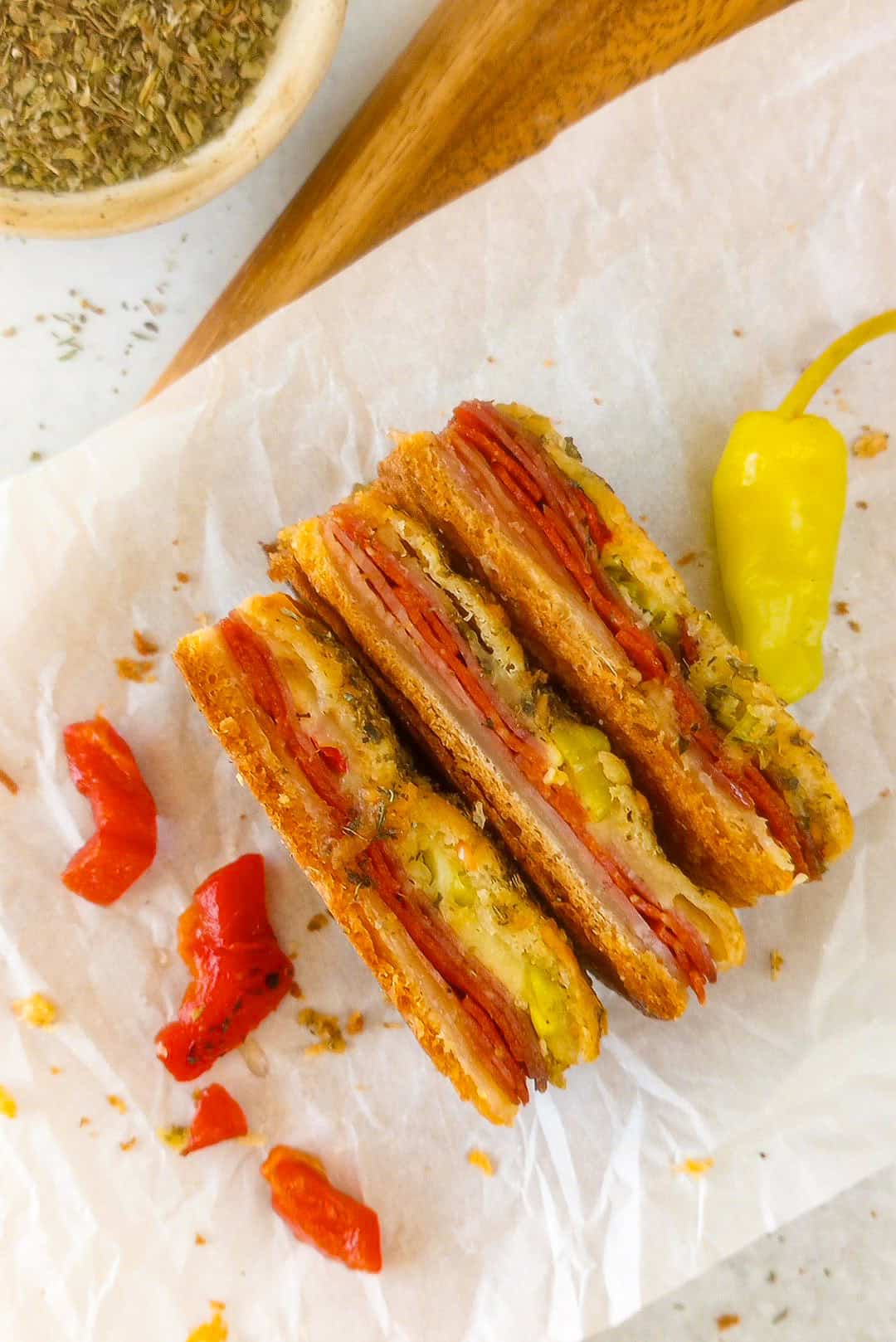 Overhead picture of antipasto squares on white parchment paper laid on the side showing meat and cheeses. Red peppers are arranged on the left side and one whole pepperoncini on the right side. Italian seasoning is in a white bowl in the upper left corner.