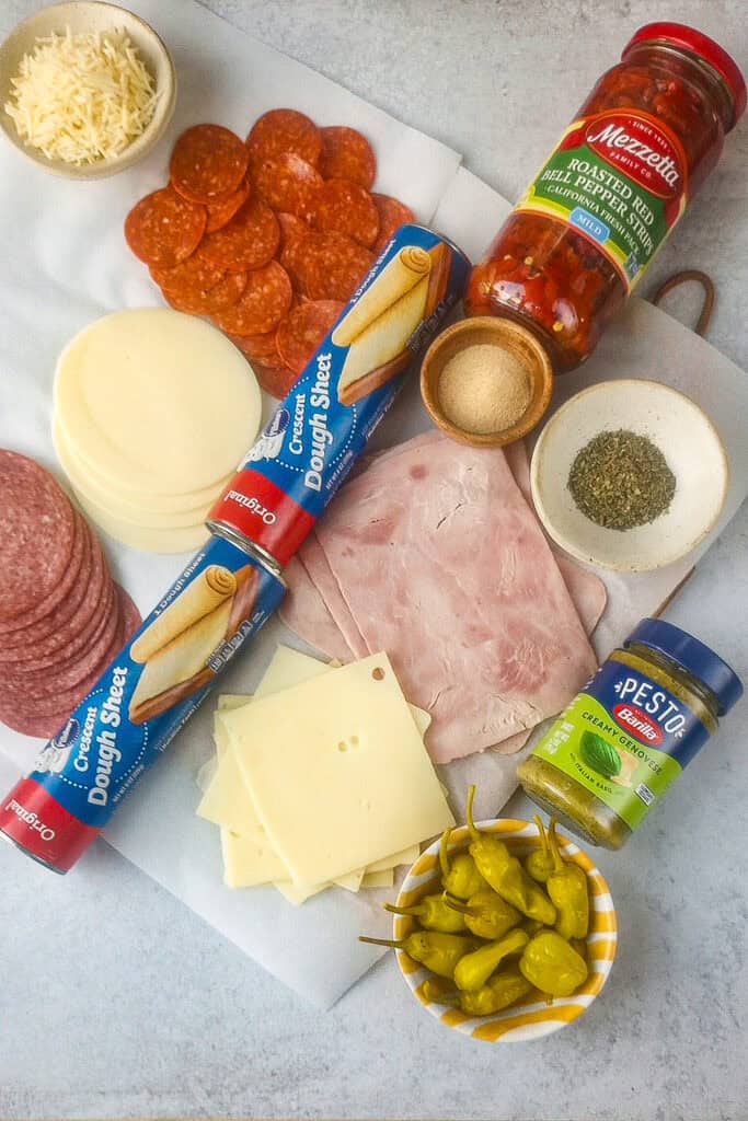 All the ingredients needed to make Italian Sub squares; Crescent roll dough sheets, Swiss cheese, ham, salami, pepperonis, pesto sauce, Italian seasoning, garlic powder, grated Parmesan cheese, Roasted red peppers, pepperoncinis, and provolone cheese. 