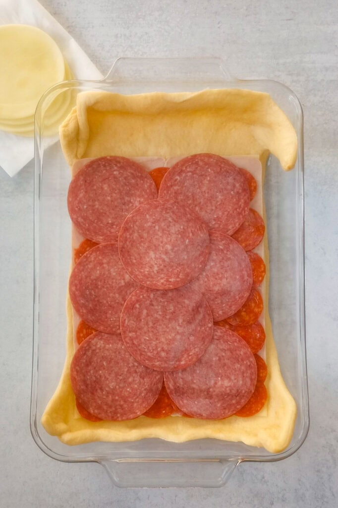 Overhead picture of crescent roll dough topped with ham, mini pepperonis, and hard salami. 