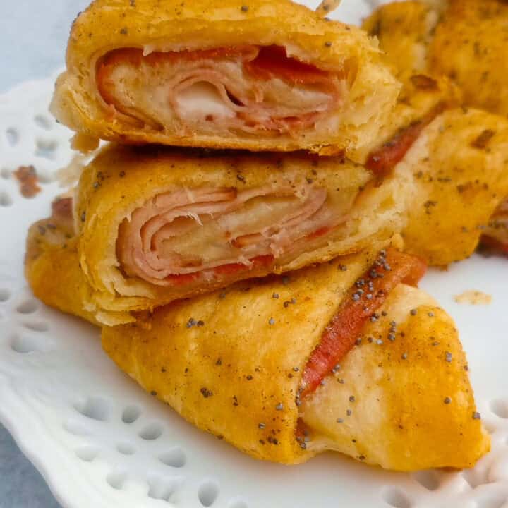 Up close picture of ham and cheese crescent rolls. One roll is sliced in half and stacked on a whole crescent roll to show the inside of bubbly, gooey, Swiss cheese and ham.